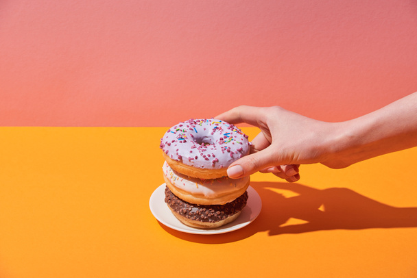 cropped view of woman taking tasty donuts on saucer on yellow desk and pink background - Fotografie, Obrázek