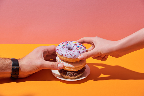 cropped view of woman and man taking tasty donuts on saucer on yellow desk and pink background - Photo, Image