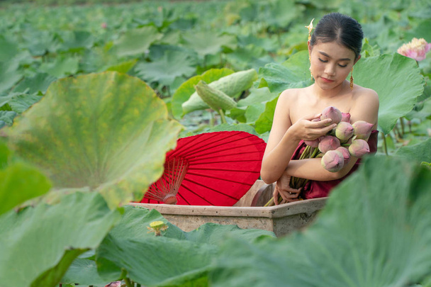 Beautiful asia women wearing traditional Thai dress and sitting on wooden boat with red paper umbrella in flower lotus lake. Her hands are holding a pink lotus. And her eyes are looking flowers. - Photo, Image