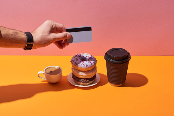 abgeschnittene Ansicht des Mannes, der nach leckeren Donuts auf Untertasse und Kaffeetassen auf gelbem Schreibtisch und rosa Hintergrund pinkelt - Foto, Bild