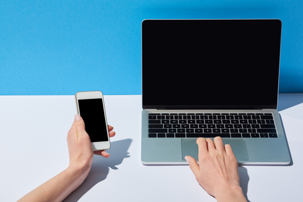 cropped view of women using laptop with blank screen and holding smartphone on white desk and blue background - Photo, Image