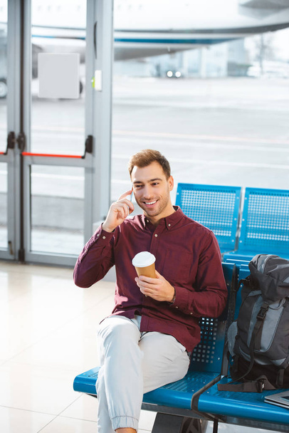 happy man talking on smartphone and holding paper cup in hand in airport  - 写真・画像