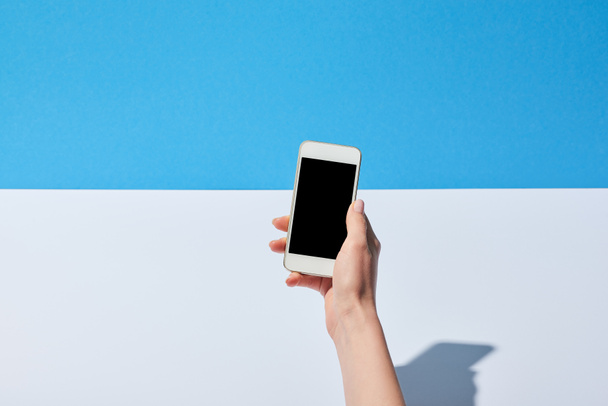 cropped view of woman using smartphone with blank screen on white desk and blue background - Foto, Imagen