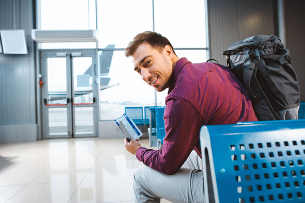 selectieve aandacht van glimlachende man die paspoort met vliegticket en zit vlak bij gate in luchthaven - Foto, afbeelding