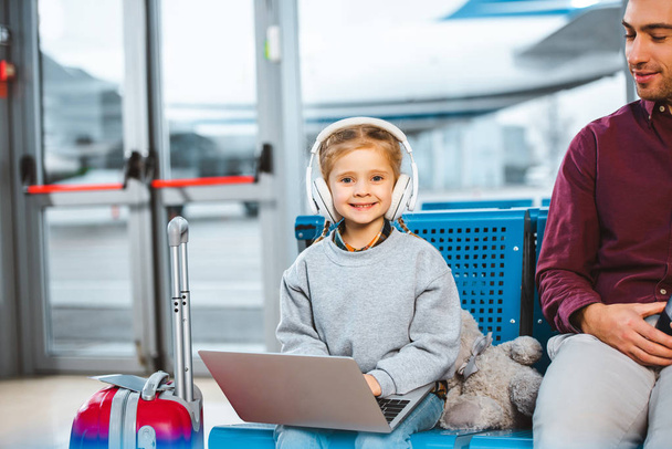 smiling kid listening music in headphones while using laptop near dad in airport  - Fotó, kép