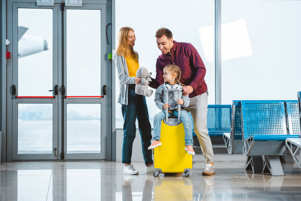 cute child sitting on suitcase while mother holding teddy bear and standing near husband in departure lounge - Photo, Image