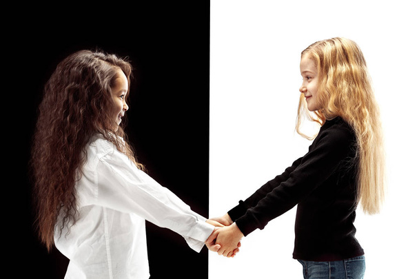 The portrait of two happy holding hands girls on a white and black studio background. Human emotions concept. Comparison. The childhood, smile, happiness, joy, kid, friendship concept - Photo, image