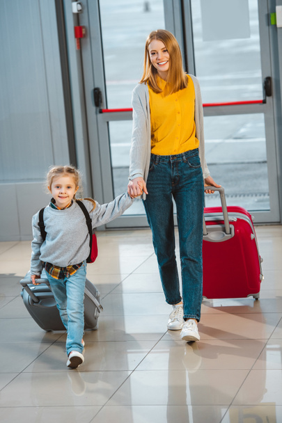 attractive mother holding hands with cute daughter and walking with baggage in airport  - Foto, Imagem