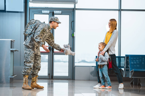 dad in military uniform giving teddy bear to daughter standing near mother in airport  - Foto, imagen