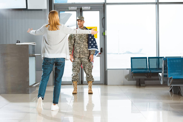 happy girlfriend with opened arms meeting boyfriend in military uniform with american flag in airport  - Zdjęcie, obraz