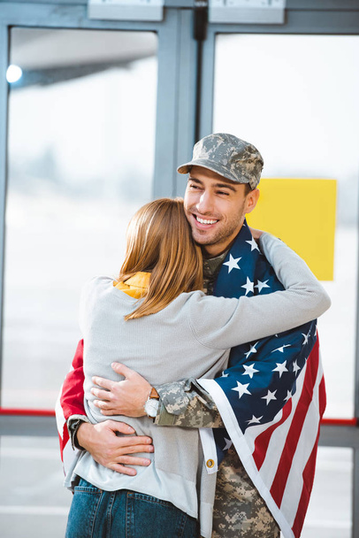 vista posteriore della donna che abbraccia il fidanzato in uniforme militare con bandiera americana in aeroporto
  - Foto, immagini