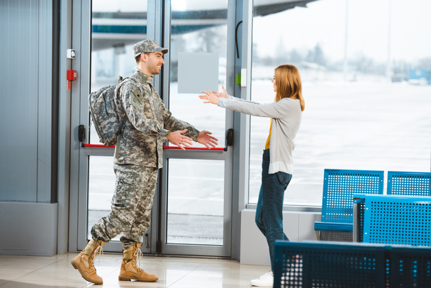 happy veteran in military uniform looking at girlfriend and standing with opened arms in airport  - Photo, Image