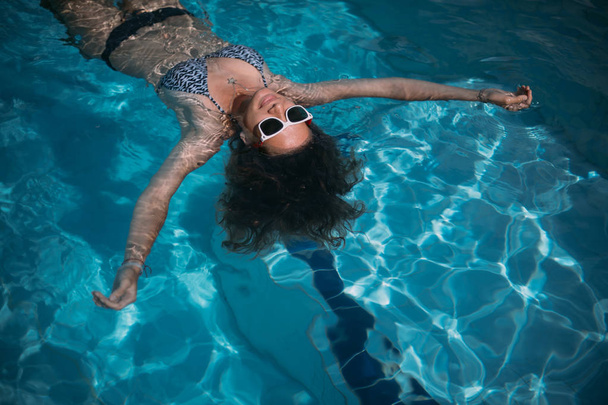 W basenie pływa młoda dziewczyna w okularach przeciwsłonecznych. Piękna kobieta odpoczywająca w wodzie w odkrytym basenie - Zdjęcie, obraz