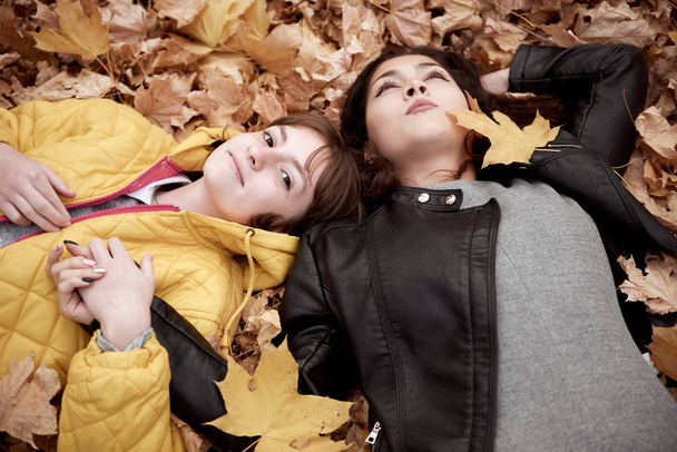 Pretty woman and teen girl are posing in autumn park. They are lying on fallen leaves. Beautiful landscape at fall season. - Photo, Image
