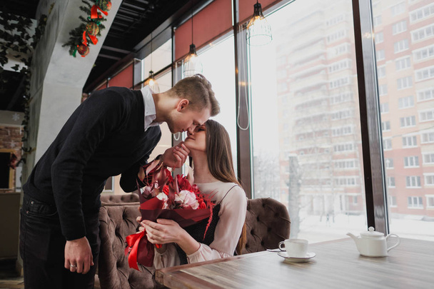 A young man gives a bouquet of red flowers to his girlfriend, wife, in a cafe by the window. - Photo, image