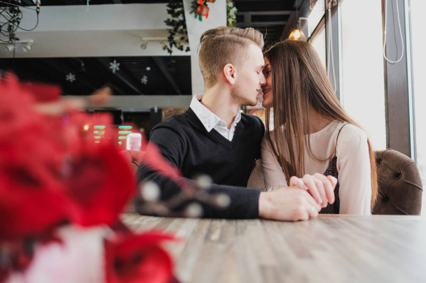 Young couple in love, man and woman on a romantic date in a cafe. Sit at a table by the window. On the table a bouquet of red flowers. - 写真・画像
