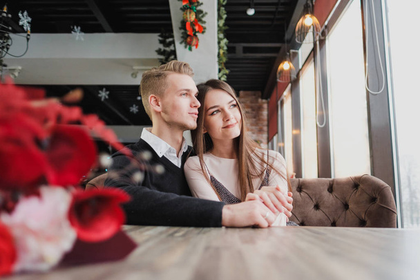 Young couple in love, man and woman on a romantic date in a cafe. Sit at a table by the window. On the table a bouquet of red flowers. - Foto, afbeelding