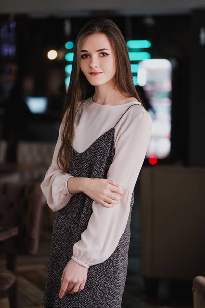 Portrait of a young girl with long hair on the background of luminous signs in the interior of a cafe or restaurant. Black dress and blouse, elegant and attractive. - Foto, Bild