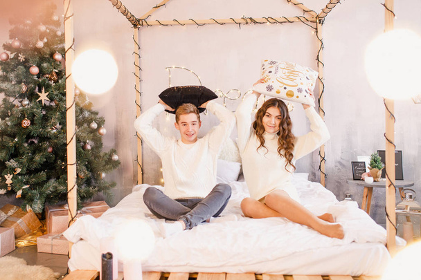 young beautiful couple are sitting on the bed on the background of the Christmas tree and smiling - Φωτογραφία, εικόνα
