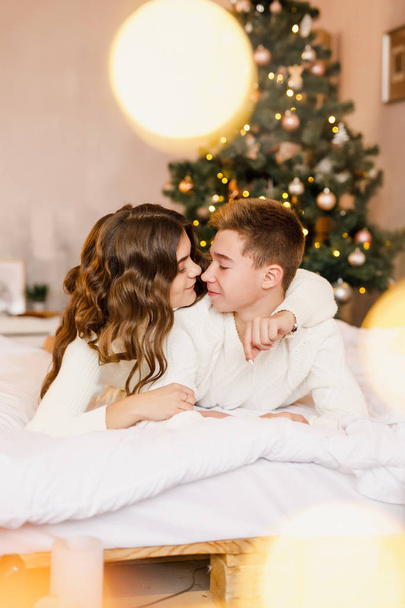 young beautiful couple are sitting on the bed on the background of the Christmas tree and smiling - Zdjęcie, obraz