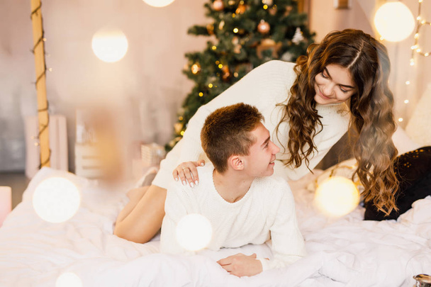 young beautiful couple are sitting on the bed on the background of the Christmas tree and smiling - Zdjęcie, obraz