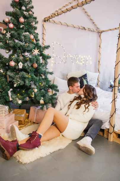 young beautiful couple are sitting on the bed on the background of the Christmas tree and smiling - 写真・画像