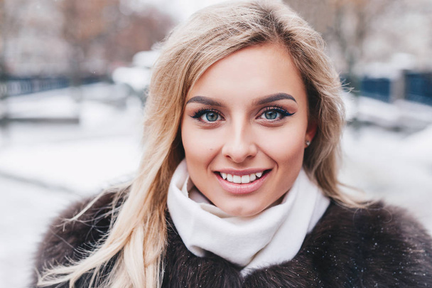 Outdoor portrait of young beautiful fashionable happy smiling girl wearing trendy fur winter coat,, posing in snow covered street. - Foto, imagen