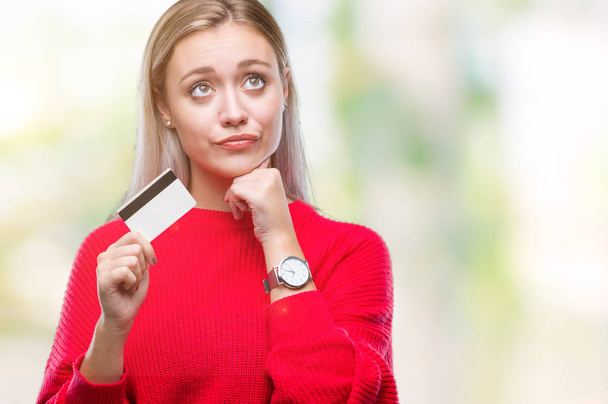Młoda blond kobieta trzymając kartę kredytową na na białym tle poważne twarz myślenia o pytanie, bardzo pomieszaną ideę - Zdjęcie, obraz
