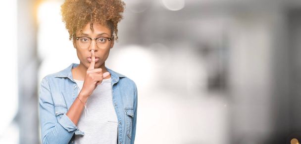 美しい若いアフリカ系アメリカ人女性の唇に指で静かにするように求めて孤立の背景にメガネを着用します。沈黙と秘密の概念. - 写真・画像