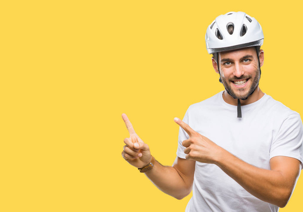 Joven hombre guapo con casco de seguridad ciclista sobre fondo aislado sonriendo y mirando a la cámara apuntando con dos manos y dedos hacia un lado
. - Foto, imagen