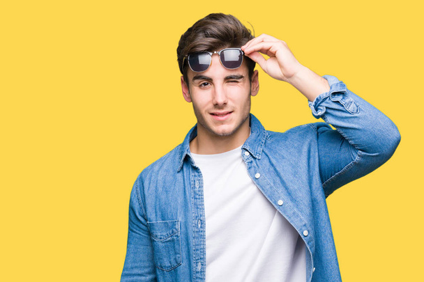Young handsome man wearing sunglasses over isolated background winking looking at the camera with sexy expression, cheerful and happy face. - Foto, afbeelding