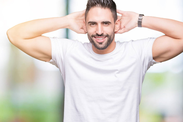 Handsome man wearing white t-shirt over outdoors background Relaxing and stretching with arms and hands behind head and neck, smiling happy - Фото, изображение