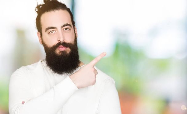 Молодой хипстер с длинными волосами и бородой в спортивной толстовке указывает пальцем в сторону, показывая рекламу, серьезное и спокойное лицо
 - Фото, изображение