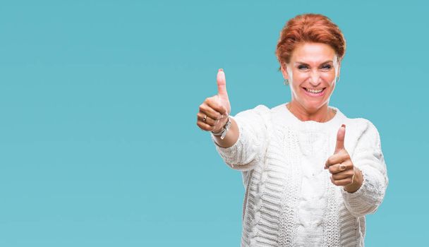 Atrractive senior caucasian redhead woman wearing winter sweater over isolated background approving doing positive gesture with hand, thumbs up smiling and happy for success. Looking at the camera, winner gesture. - Photo, image