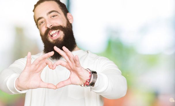 Young man with long hair and beard wearing sporty sweatshirt smiling in love showing heart symbol and shape with hands. Romantic concept. - Fotó, kép