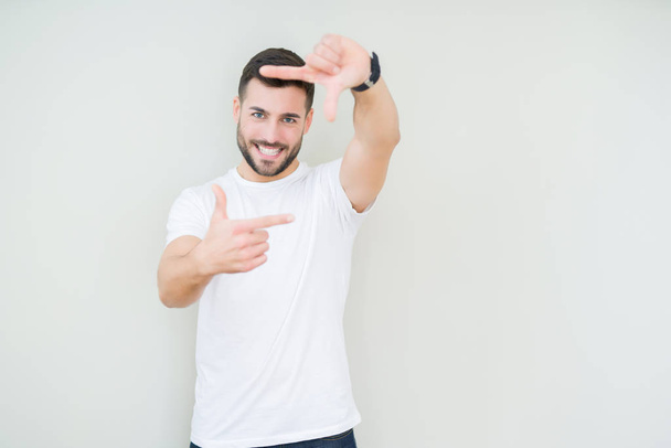 Mladý pohledný muž nosí ležérní bílé tričko izolované pozadí provedení rámu s rukama a prsty s šťastné tváře. Koncept kreativity a Fotografie. - Fotografie, Obrázek