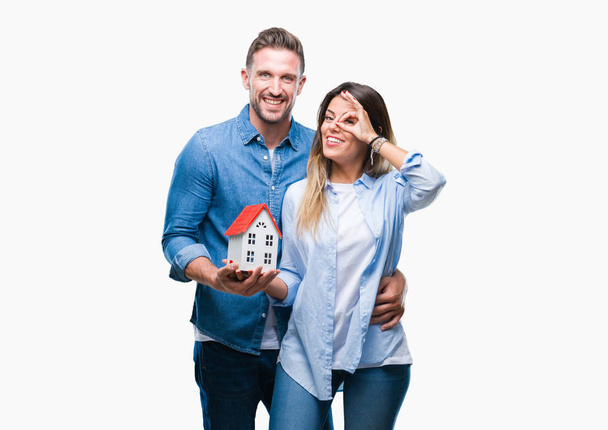 Young couple in love holding house over isolated background with happy face smiling doing ok sign with hand on eye looking through fingers - Photo, image