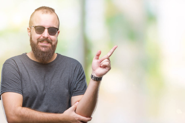 Young caucasian hipster man wearing sunglasses over isolated background with a big smile on face, pointing with hand and finger to the side looking at the camera. - Fotoğraf, Görsel