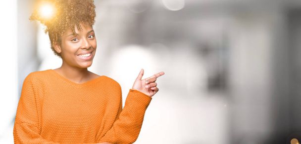 Belle jeune femme afro-américaine sur fond isolé avec un grand sourire sur le visage, pointant la main et le doigt vers le côté en regardant la caméra
. - Photo, image