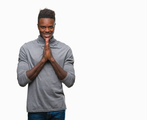 Молодой африканский американец на изолированном фоне молится руками, прося прощения, уверенно улыбаясь.
. - Фото, изображение