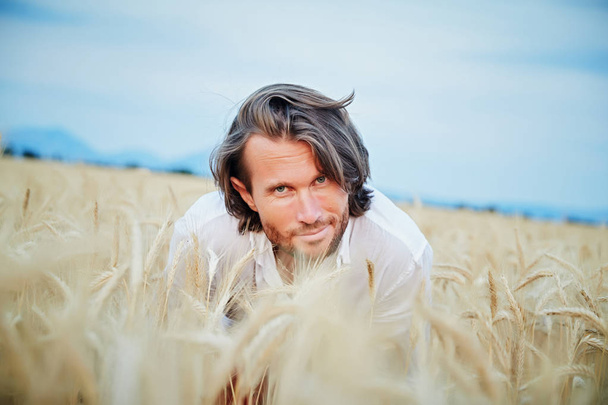 The beautiful brutal young man with long brunette hairs poses in the field with wheat, suntanned man, easy beard, sunset light, turns, smiles, flirts, happy, white shirt, Red shorts, rye field - Fotoğraf, Görsel