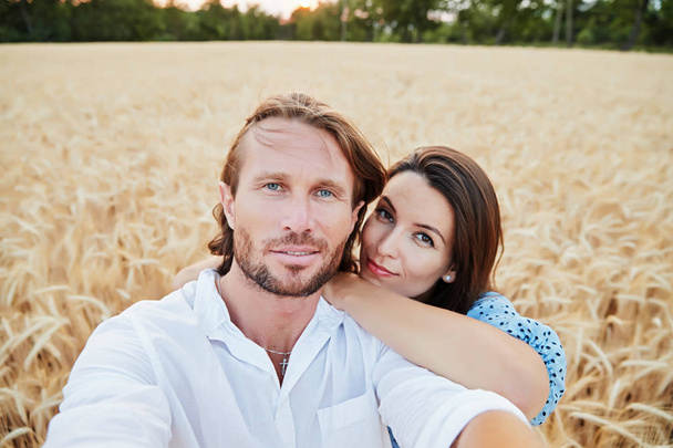 The beautiful couple embraces in the field of a rye and looks in a camera, the beautiful eyes, happy smiles, the long hair, an easy beard, wheat ears on background,  blue dress and white shirt - Photo, image