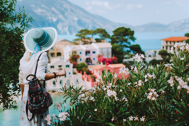 Elegant woman with straw hat and white clothes enjoying view of colorful village Assos on sunny day. Stylish female visiting Kefalonia in summer time on Greece travel vacation. - Foto, imagen