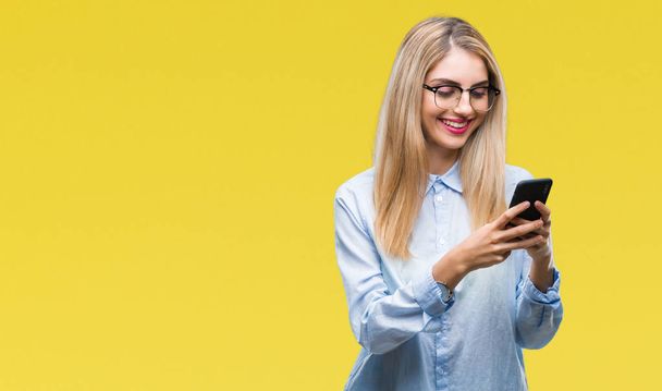 Genç güzel sarışın iş kadını ayakta ve diş gösterilen kendine güvenen bir gülümsemeyle gülümseyerek mutlu bir yüz ile izole arka plan üzerinde Smartphone kullanarak - Fotoğraf, Görsel