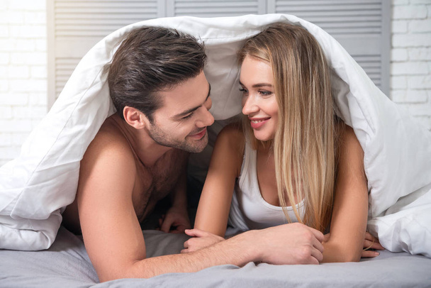 portrait of happy young couple looking at each other lying under the blanket on the bed at home - Fotografie, Obrázek