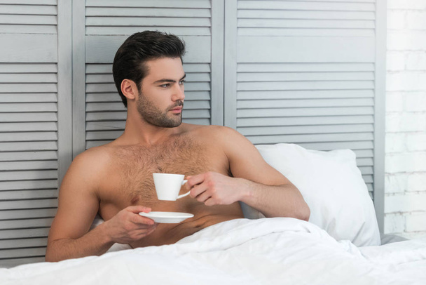handsome man drinking coffee in the bed, looking away - 写真・画像