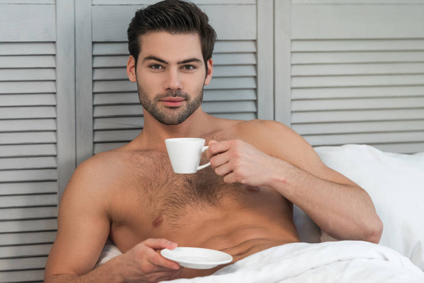 retrato de un joven desnudo bebiendo café en la cama y mirando a la cámara
 - Foto, Imagen