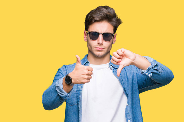 Young handsome man wearing sunglasses over isolated background Doing thumbs up and down, disagreement and agreement expression. Crazy conflict - Foto, imagen