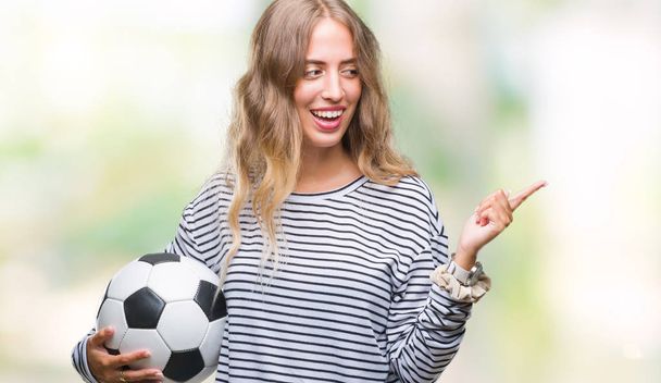 Piękna młoda kobieta blonde holding piłka nożna Piłka nożna na białym tle bardzo szczęśliwy, wskazując z dłoni i palców z boku - Zdjęcie, obraz