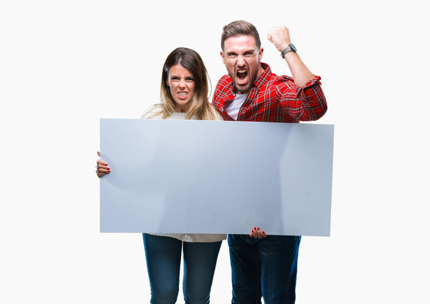 Young couple together holding blank banner over isolated background annoyed and frustrated shouting with anger, crazy and yelling with raised hand, anger concept - Zdjęcie, obraz